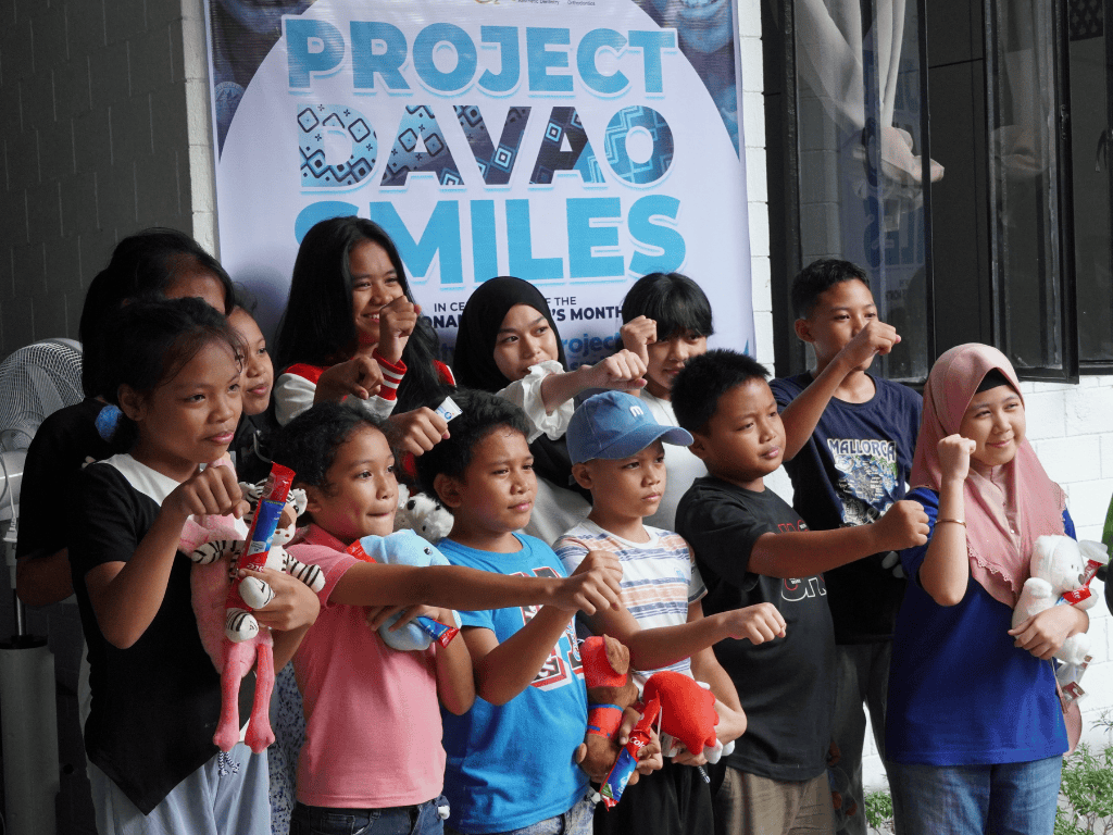 A group of children, standing in front of the Davao Smiles Poster, smiling and holding up their fists like a fist-bump gesture