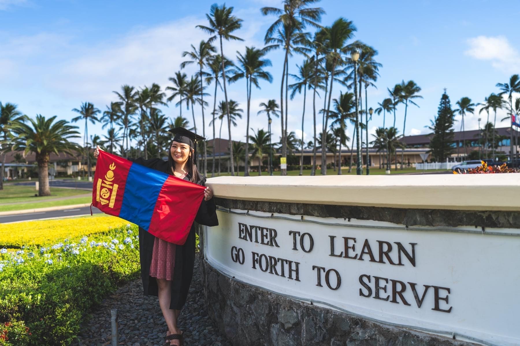 Marla Chinbold in front of a sign saying "enter to learn, go forth to serve" and holding the Mongolian flag while wearing a cap and gown.